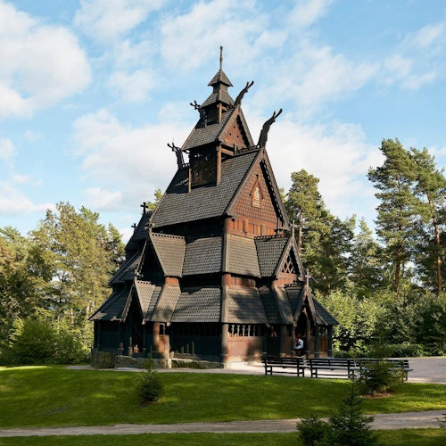 Деревянная столбовая церковь с замысловатым дизайном крыши, окружённая пышной зеленью и деревьями, под частично облачным небом.