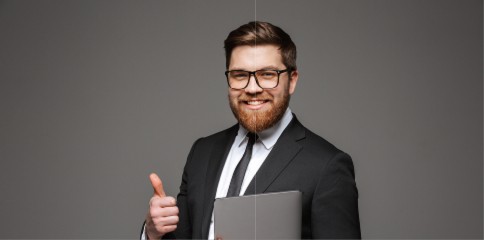 A man with a beard, wearing a black suit and tie, smiles confidently while holding a gray laptop and giving a thumbs-up against a plain gray background, conveying professionalism and positivity.