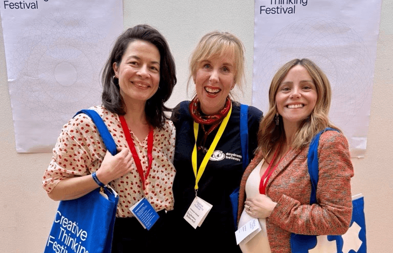Three people stand side by side at an indoor event, each wearing a lanyard and holding branded tote bags. Posters reading “Creative Thinking Festival” are displayed on the wall behind them, and the group is standing on a wooden floor against a light‑coloured background.