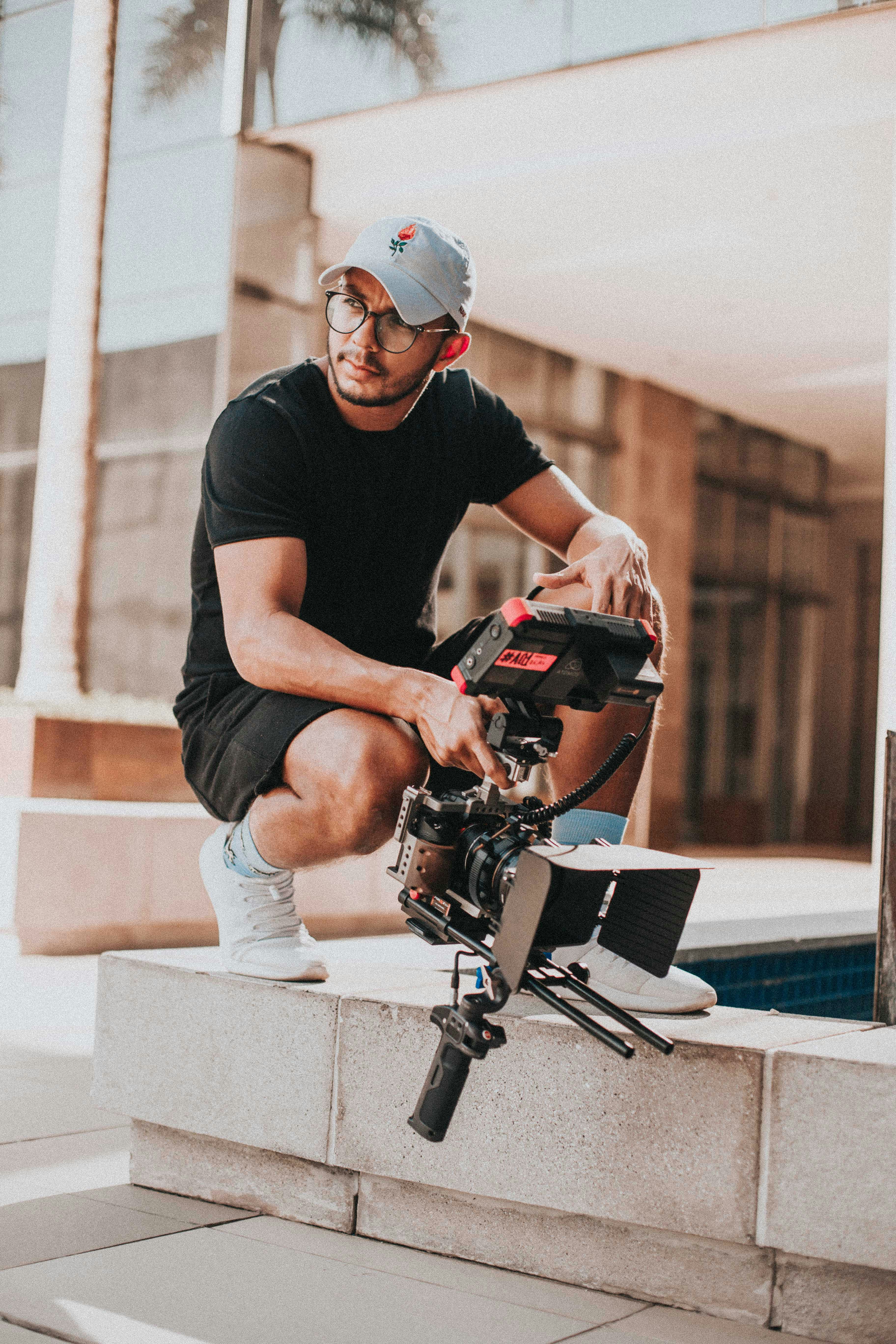 A person in casual attire crouches on a stone ledge, holding a professional video camera, with a modern architectural backdrop and a palm tree visible in the background.