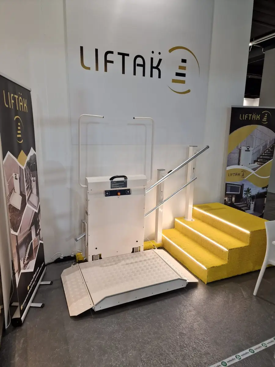 Liftak incline platform stairlift on display at a trade show stand, shown with platform extended on a small staircase in front of Liftak branded signage