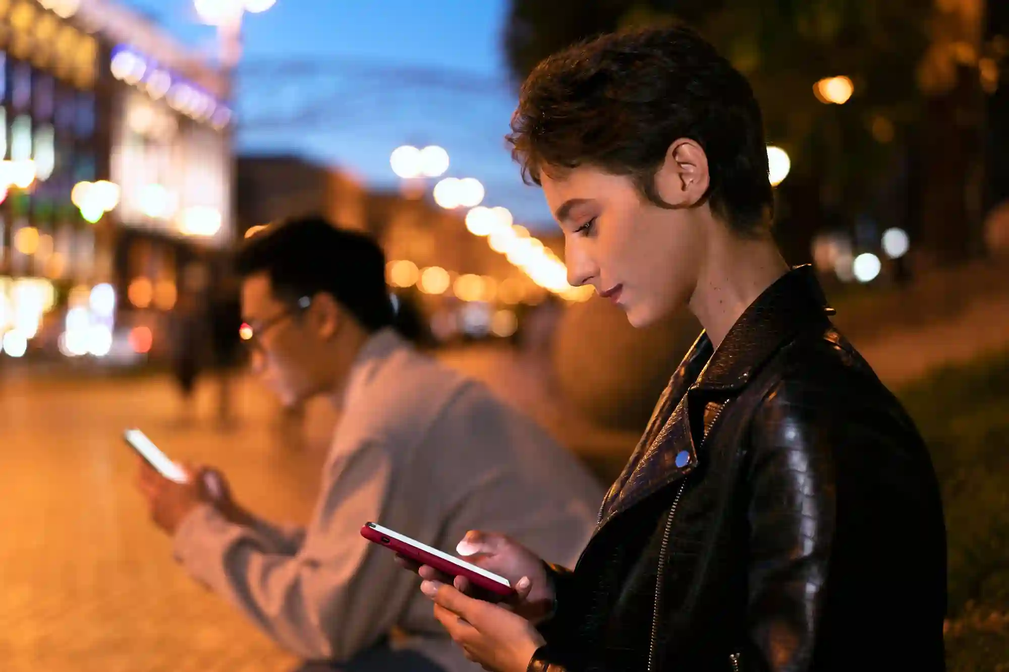 Two people sit outdoors at night, focused on their illuminated smartphones against a blurred city background.