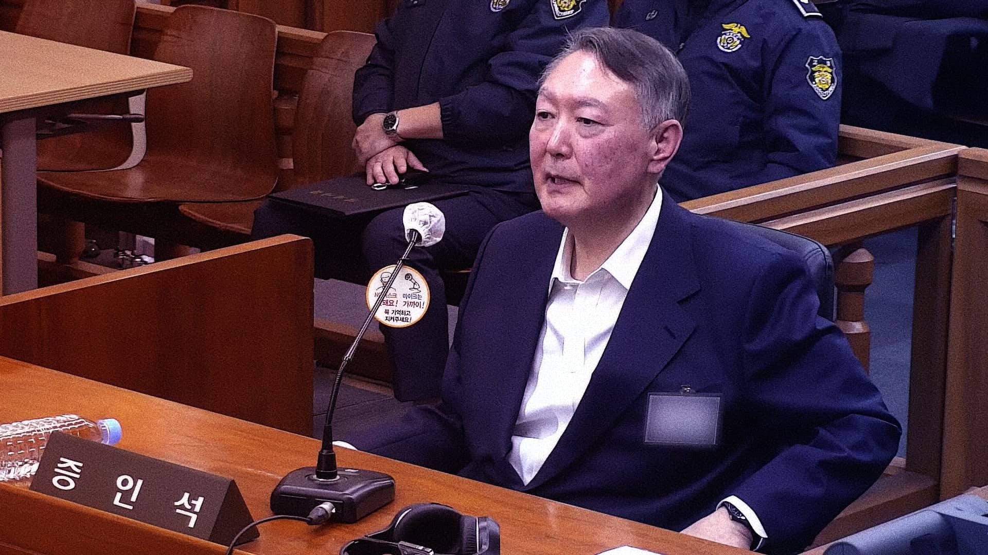 Cựu Tổng thống Hàn Quốc Yoon Seok Yeol ngồi tại vị trí bị cáo trong phiên tòa xét xử vụ nội loạn, mặc vest xanh navy với áo sơ mi trắng, phía sau là cảnh sát tư pháp