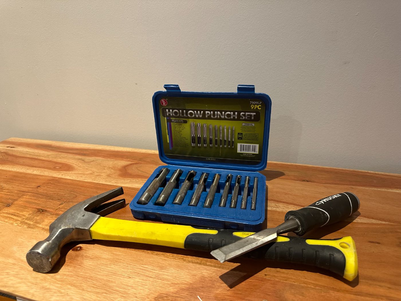 Ensemble d'outils comprenant un marteau à manche jaune, un ciseau à bois Husky et une boîte bleue de poinçons creux sur une table en bois