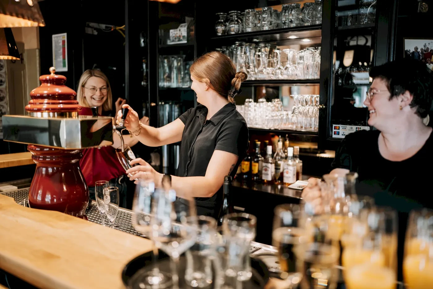 Eine Barkeeperin zapft Bier in einem Restaurant. Zwei Frauen sitzen am Tresen und unterhalten sich.