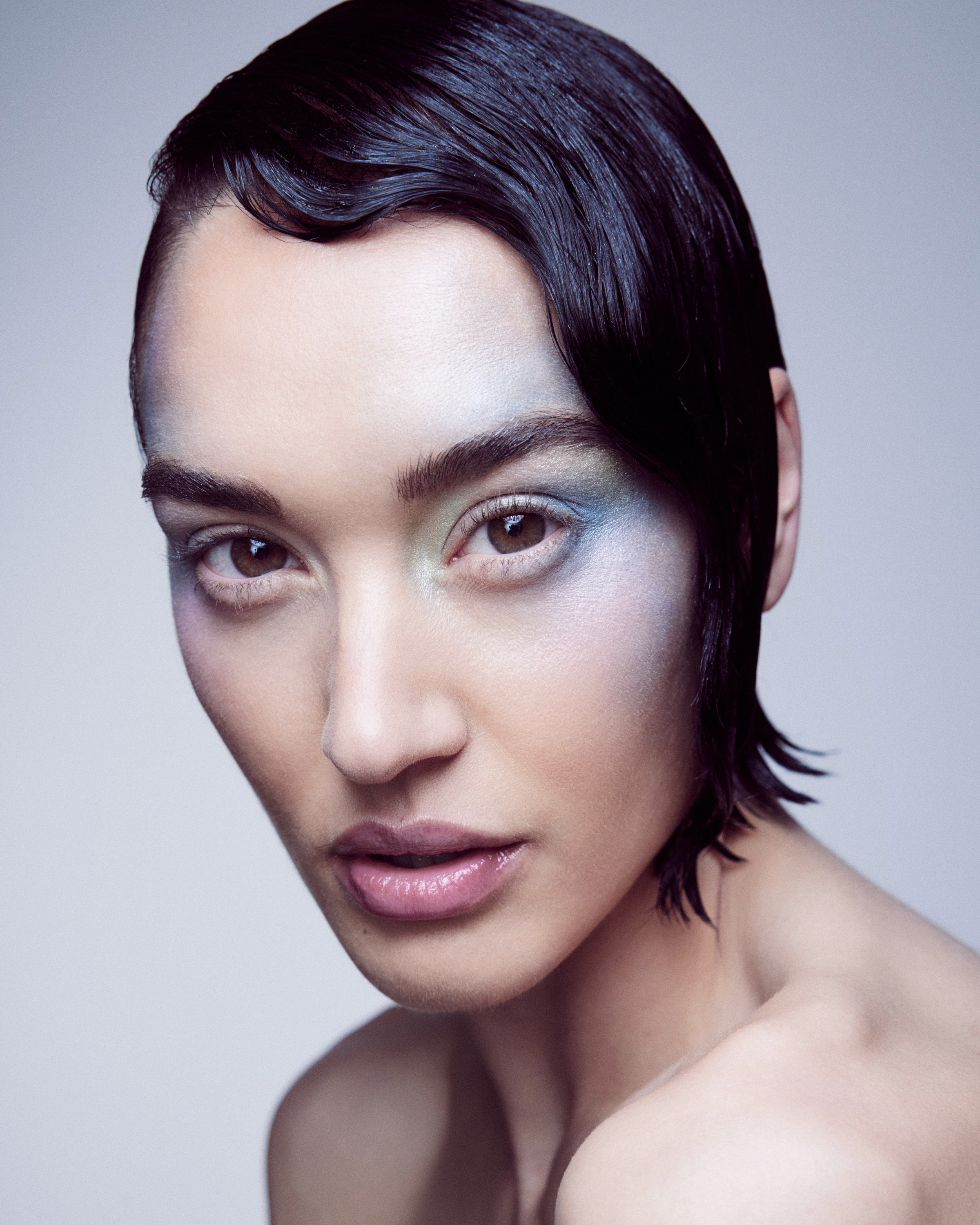 A female model with slicked black hair with a wavy fringe and satin silver makeup on her outer eye and cheek bone.