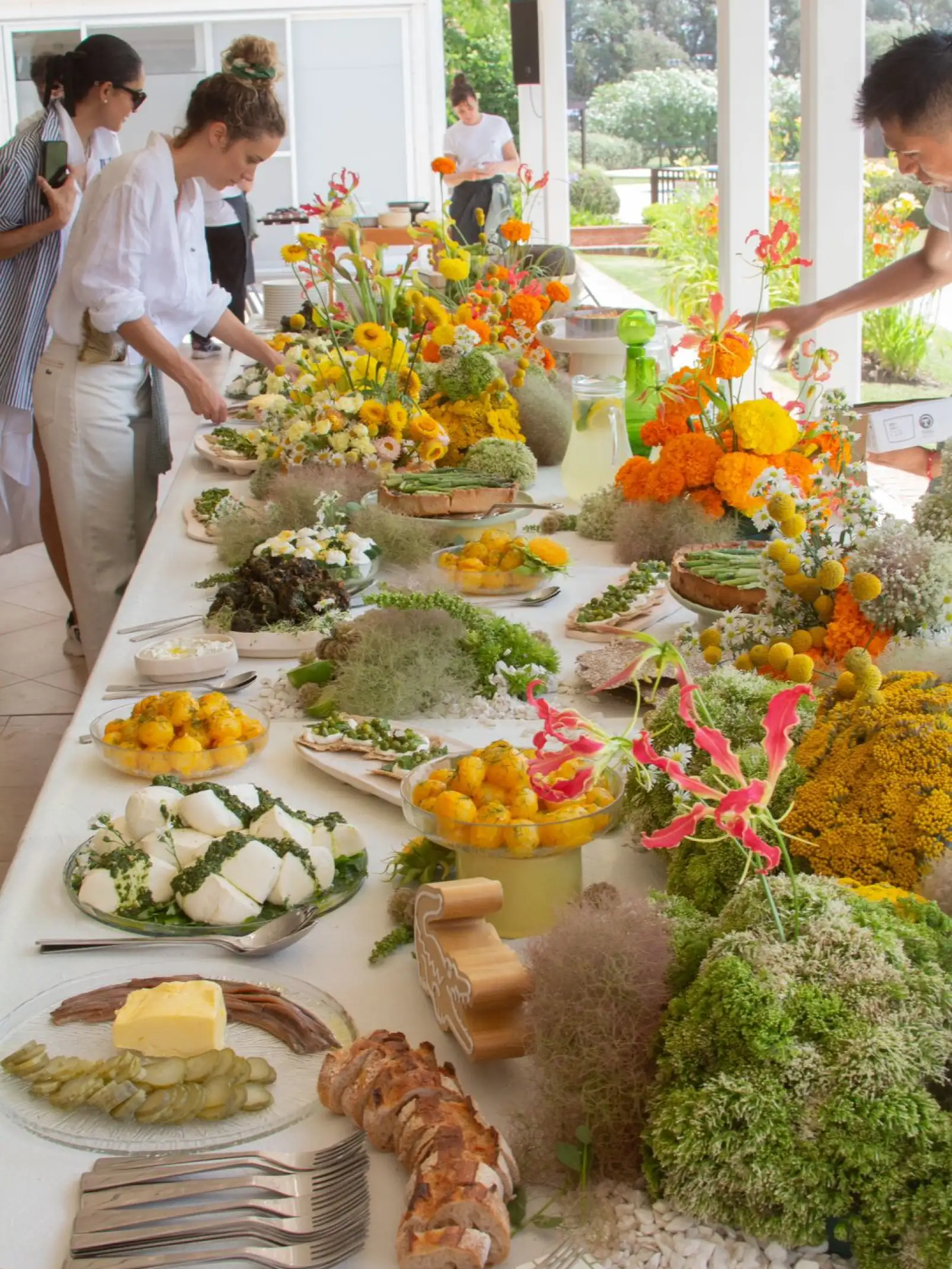 Mesa buffet decorada con arreglos florales en tonos amarillos, naranjas y verdes, acompañada de platos con quesos, panes, vegetales y frutas frescas, mientras invitados se sirven en un ambiente al aire libre.