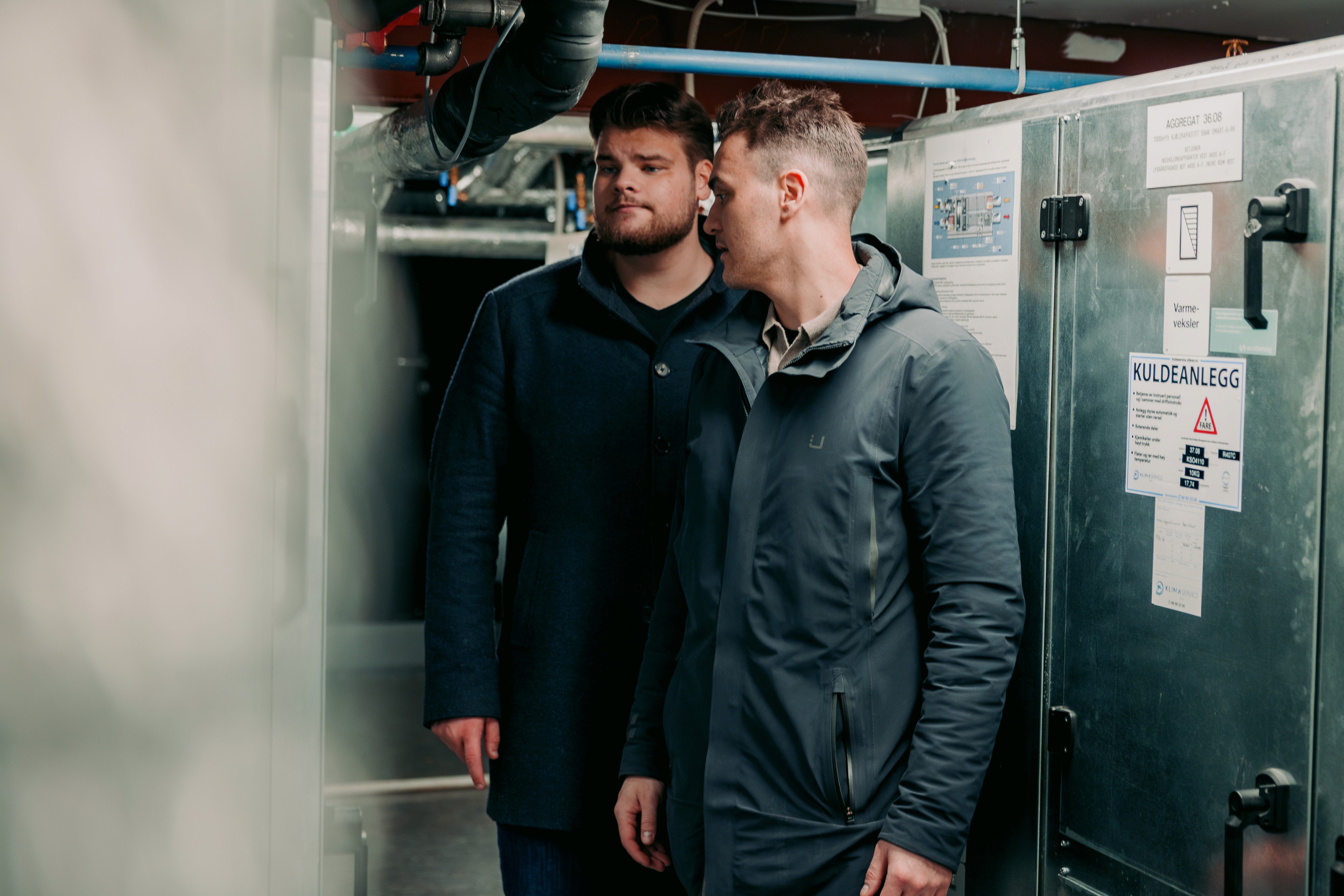 Two men in a dark utility room examine a control panel marked "Kullanlegg," with visible pipes and machinery in the background.