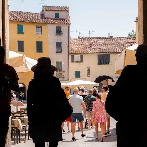 People walking through a Mediterranean street with old buildings, outdoor tables, and umbrellas, viewed from a shaded passage.