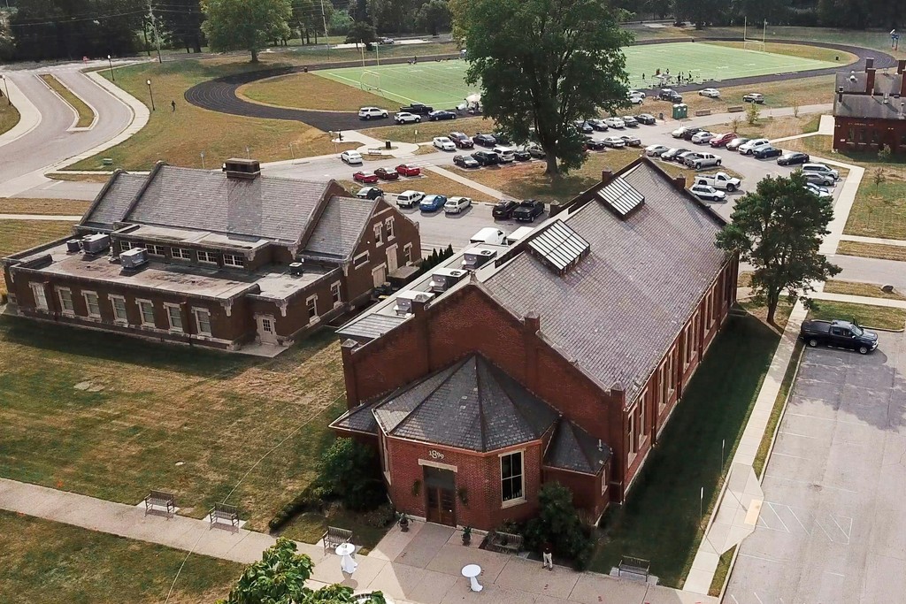 Drone view of a historic brick event venue with a turret entrance, guest parking, and open green grounds on a summer day