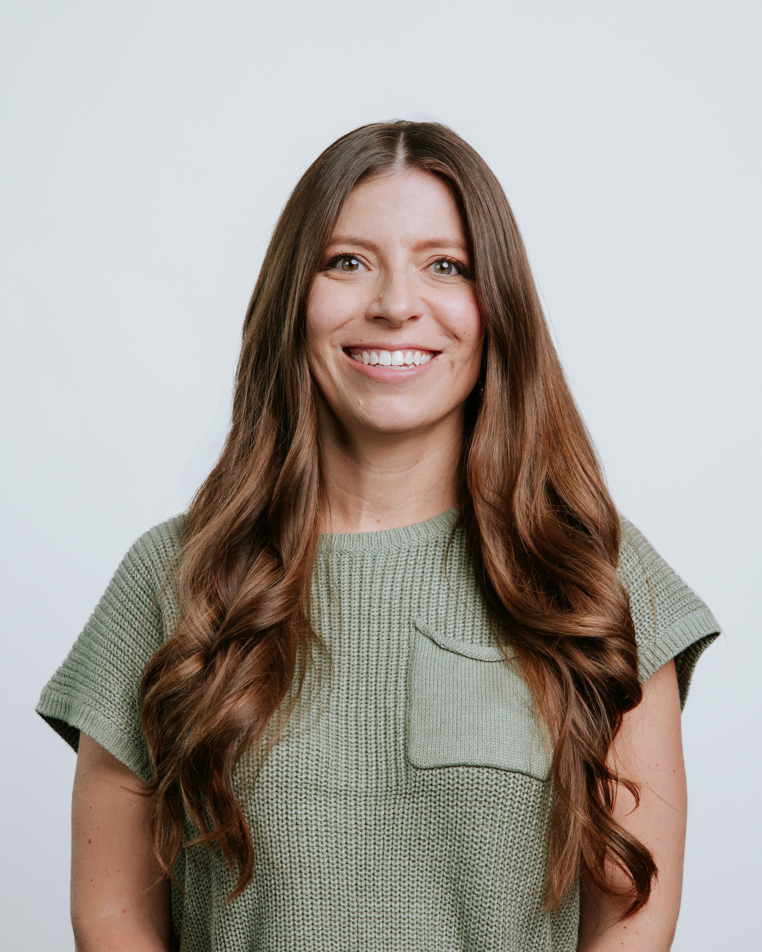 A smiling person with long, wavy brown hair, wearing a sleeveless white top, against a light background.