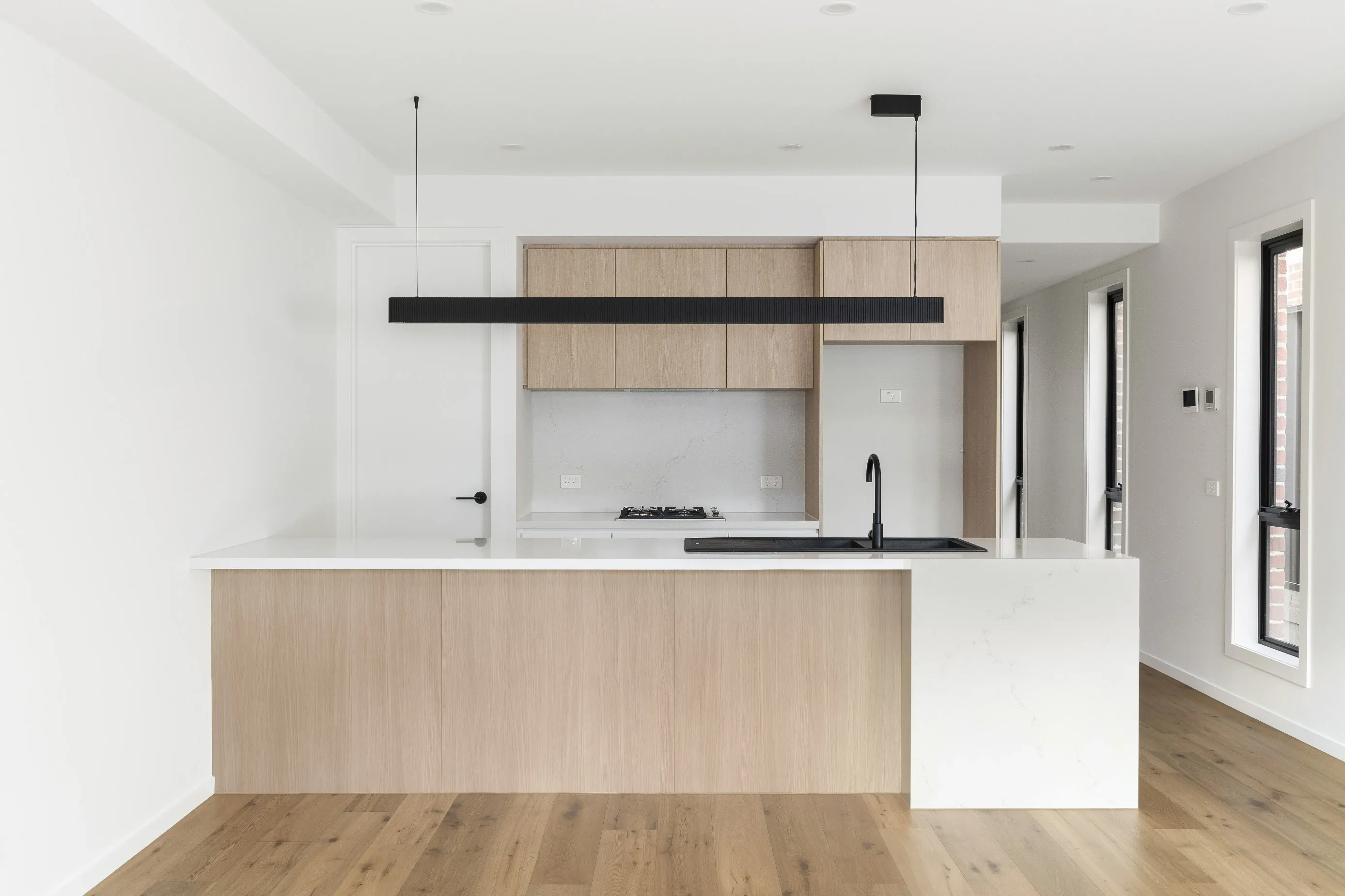 Open-plan kitchen and living with white walls, oak floors, pale-oak cabinetry, a black fluted linear pendant and a white-and-oak island with black gooseneck tap and sink, beside black-framed windows