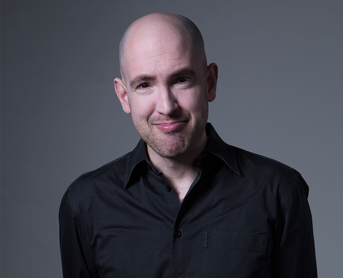 The image shows a man with a shaved head and a short beard or stubble. He is wearing a black button-down shirt and looking directly at the camera. The background is a solid gray color. The man is positioned centrally in the frame and is the sole focus of the image. No other objects or text are visible in the image.