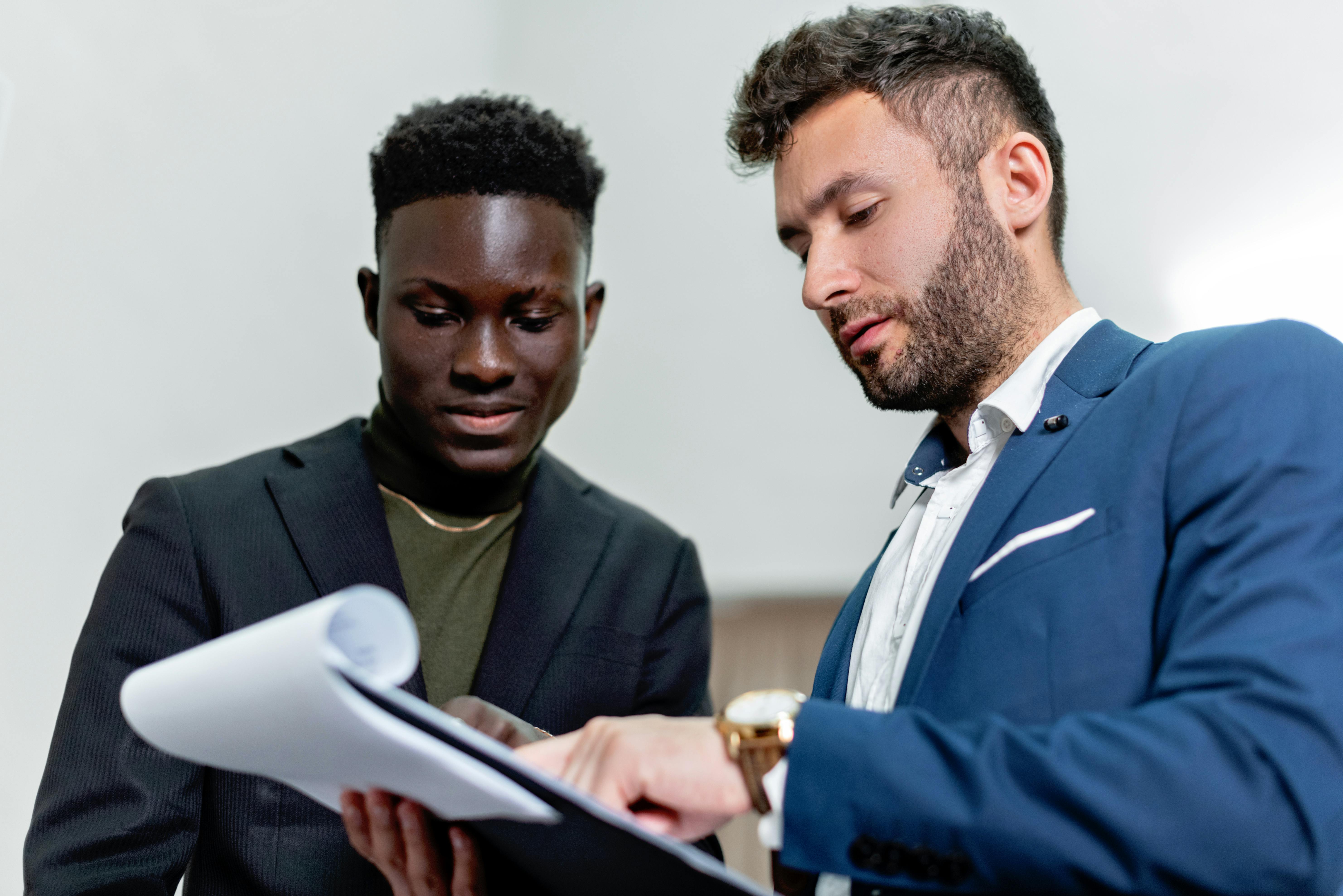 Two business professionals, one wearing a black suit and the other in a blue suit, discuss documents together.