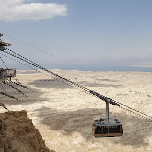 A cable car suspended mid-air over a desert landscape, connecting to a cliffside platform with a distant body of water in the background.