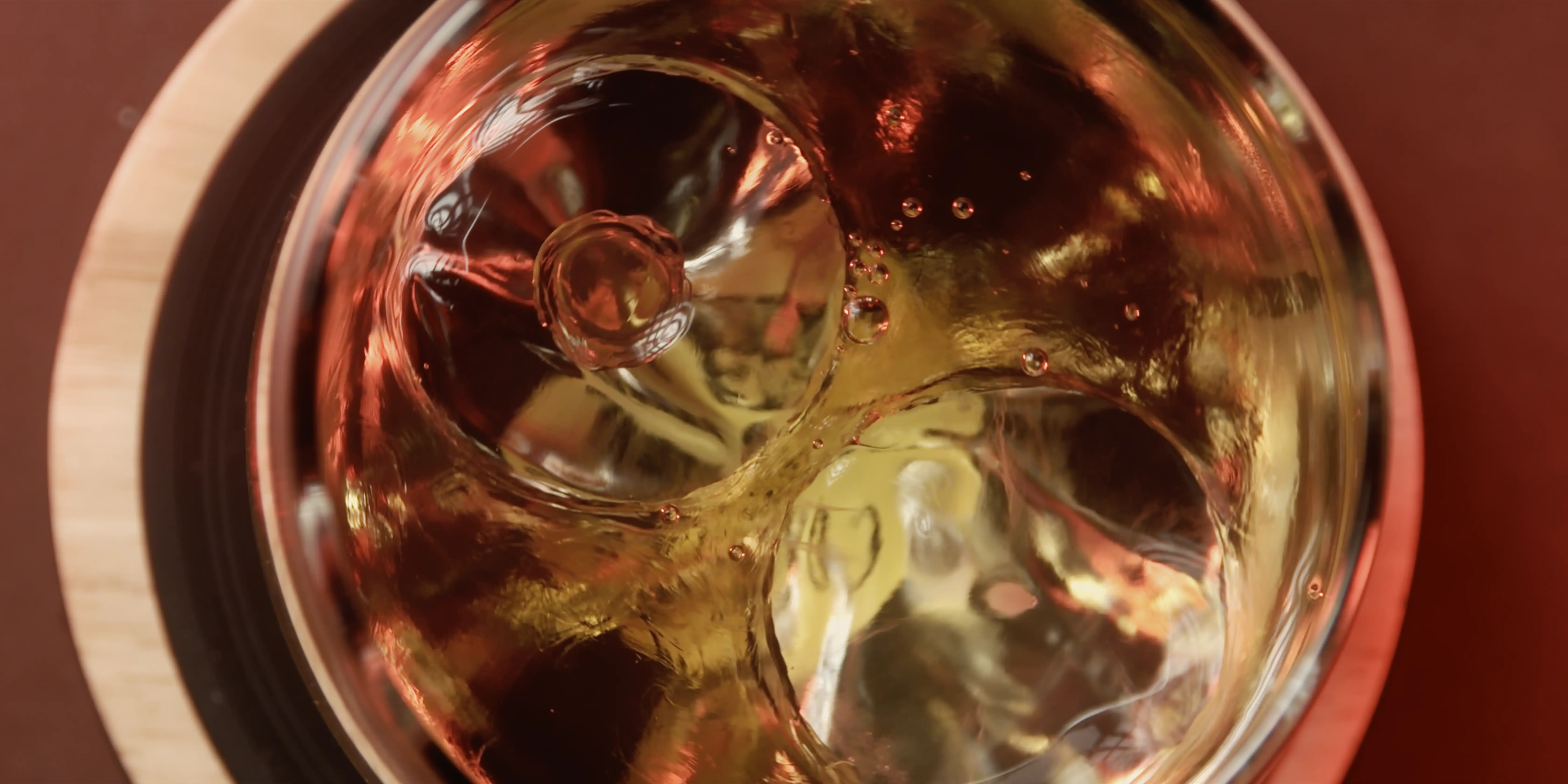 A glass filled with a fizzy, amber-colored beverage is placed on a wooden coaster, showing bubbles rising to the surface and reflecting warm light, creating a refreshing, inviting appearance.