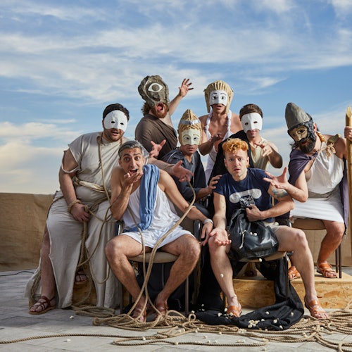 一群戴着各种面具、身穿不同服装的人在以蓝天为背景的舞台上摆出富有戏剧感的姿势。