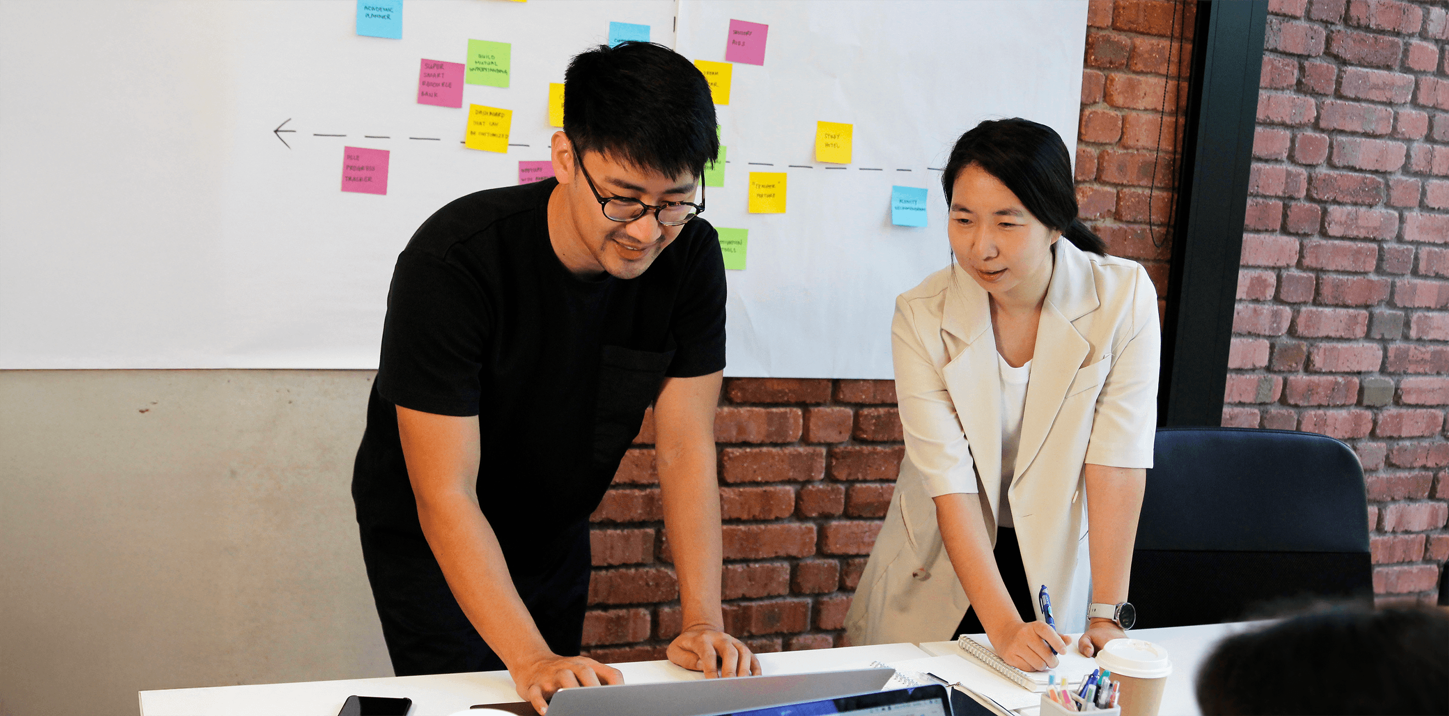 Two members of 55 Minutes collaborating in a modern workspace, discussing ideas in front of a laptop and a board with sticky notes.