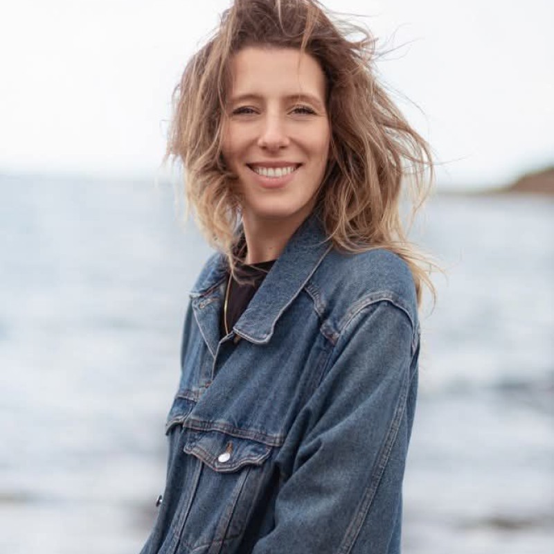 A person with curly hair and glasses smiles at the camera, seated in a cozy workspace with warm lighting.
