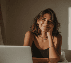 A woman sitting at a desk, smiling while resting her head on her hand, with a laptop in front of her.