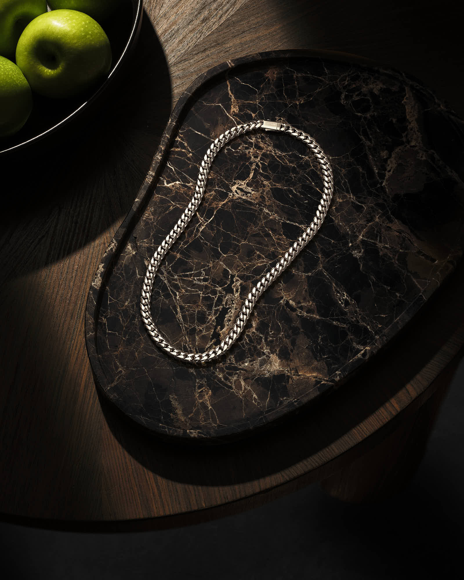 Silver chain necklace on a dark marble tray with green apples.