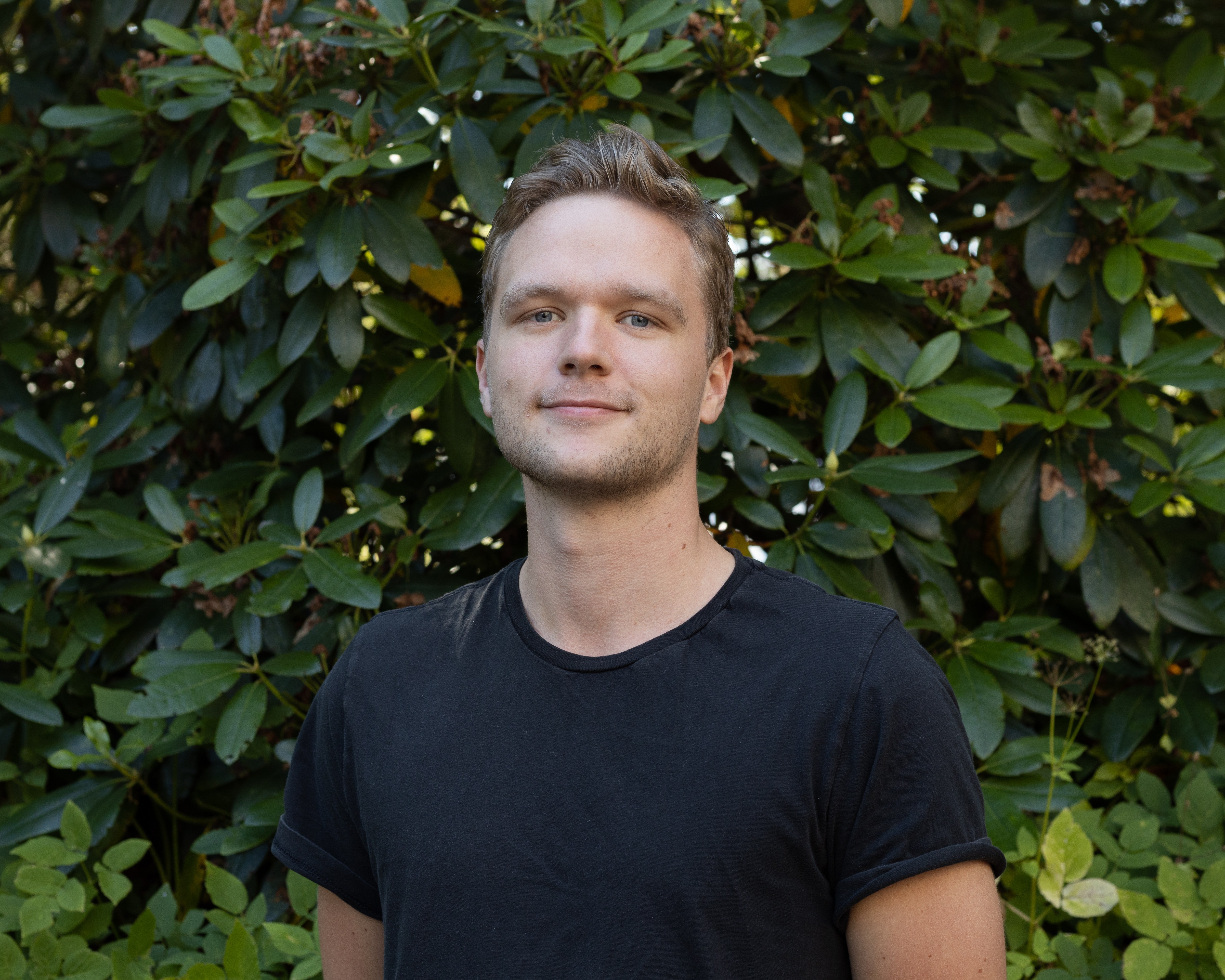 A smiling young man with crossed arms, wearing a plaid shirt and white t-shirt, poses against a dark background.