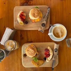 Top-down view of a wooden table with two breakfast sandwiches on wooden boards, served with strawberries and a small salad, alongside a cup of latte, iced drink, and utensils.
