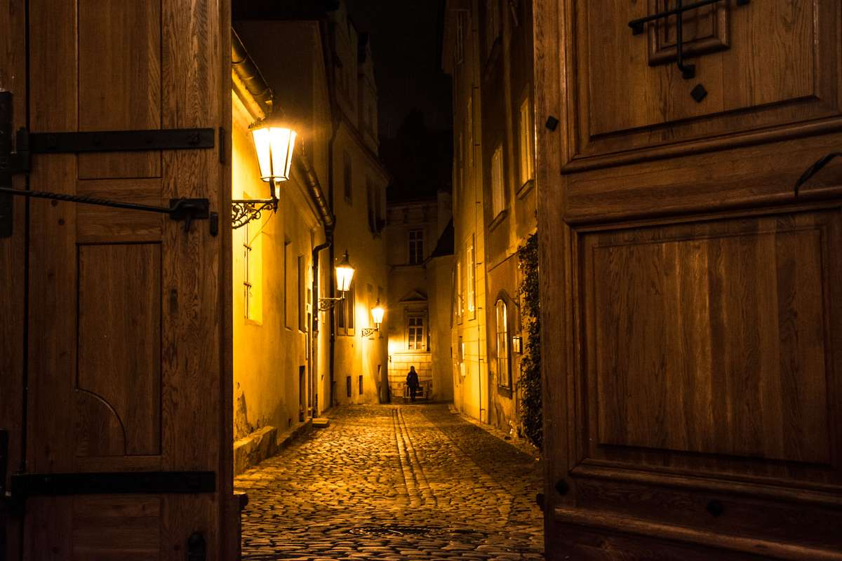 A narrow cobblestone alleyway is illuminated by glowing lanterns at night between two large wooden doors and aged stone walls.