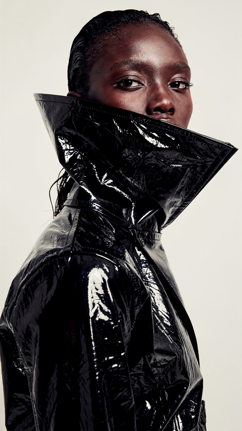 A person with slicked-back hair wears a glossy black trench coat with a high collar, looking serious and intense against a neutral background.