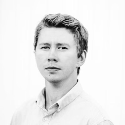 A person with medium-length brown hair wearing a dark, textured coat and a burgundy turtleneck. They have a calm, neutral expression.