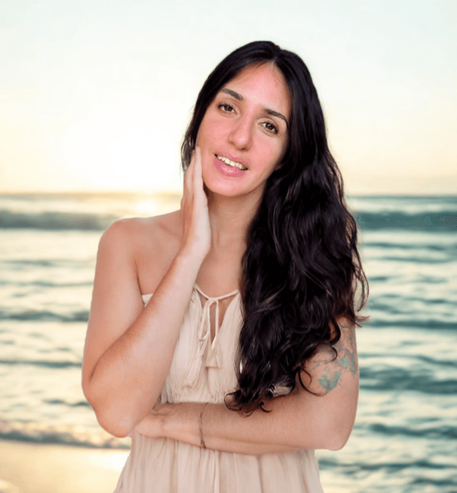 A woman wearing a boho dress is standing her back to the ocean at sunset. She has her hand in her neck, and looks confident in her body and feminine