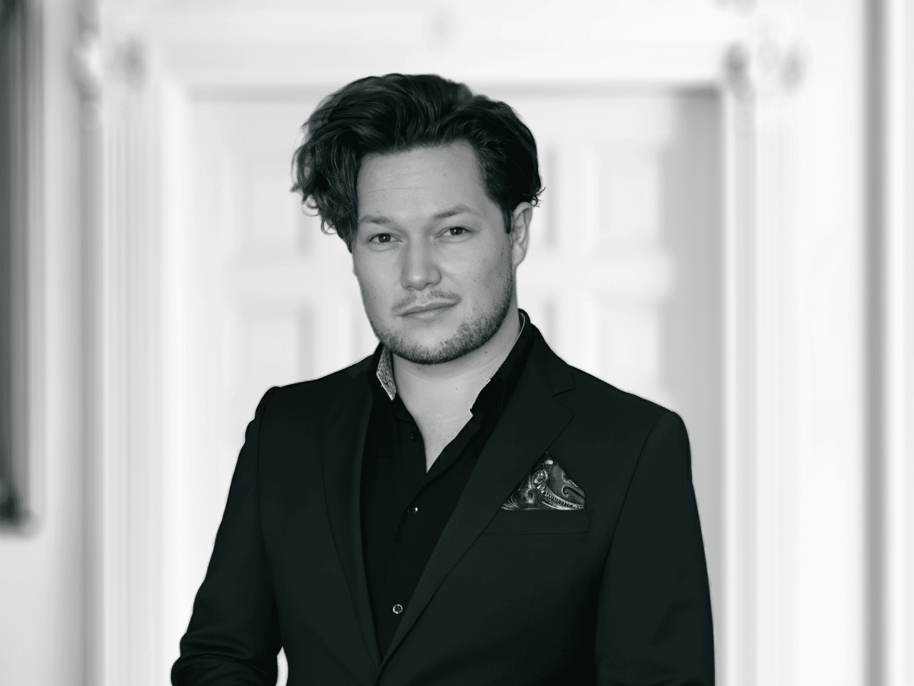 Porträt eines Mannes mit dunklem, voluminösem zur Seite gekämmtem Haar. Er trägt ein schwarzes Sakko über einem dunklen Hemd und blickt freundlich in die Kamera. Der Hintergrund ist grün und zeigt einen stilisierten Fennek-Kopf in weiß..