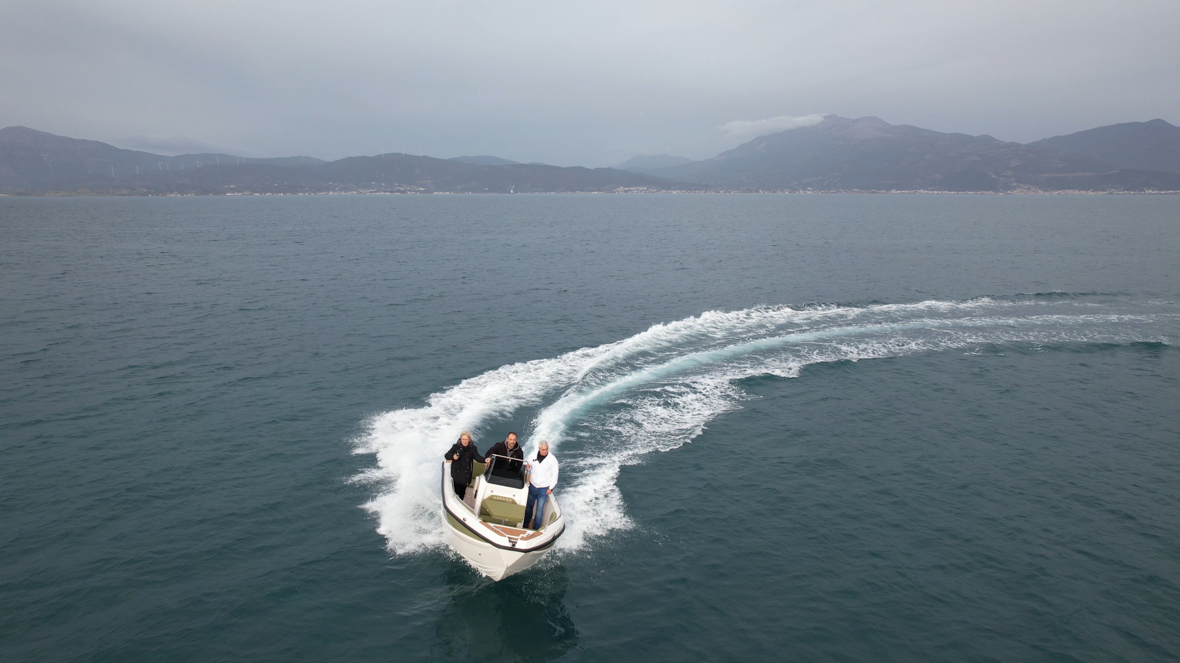 White speedboat with passengers making a sharp turn in turquoise waters, creating dramatic wake trails with Cycladic mountains in the background.