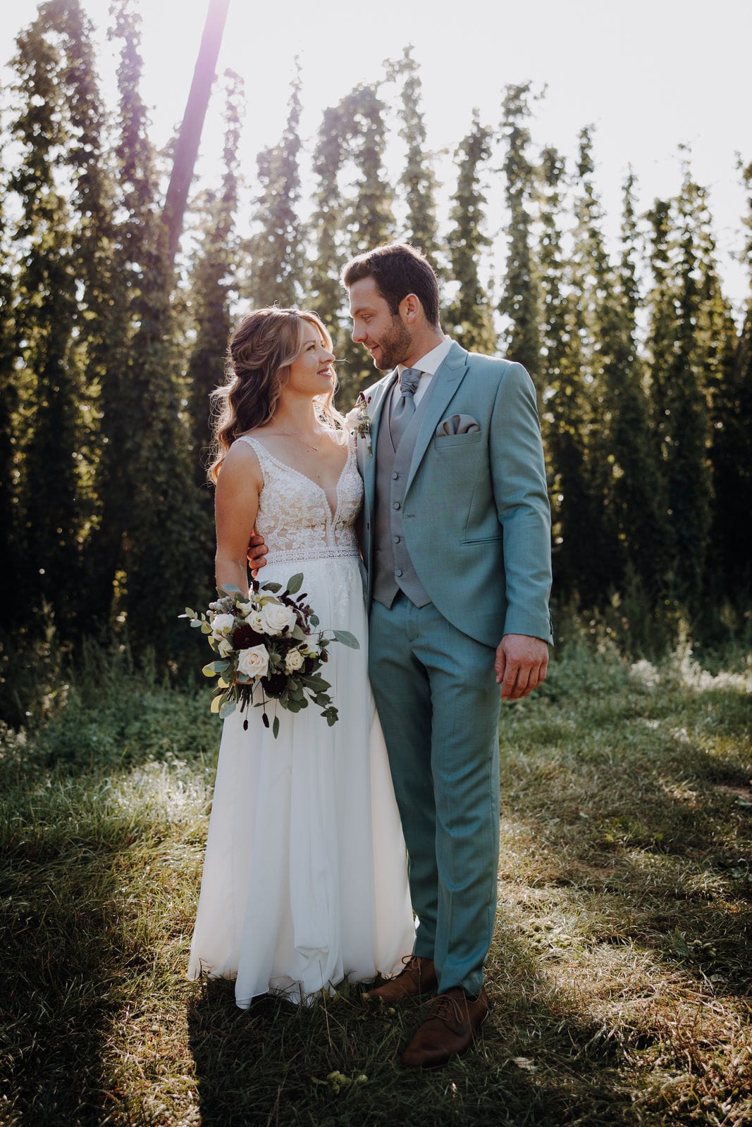 Kundenfeedback Hochzeitspaar für Maria Wiethaler freie Rednerin und Sängerin. Romantisches Brautpaar im Sonnenlicht: Die Braut trägt ein zartes Spitzen-Hochzeitskleid mit fließendem Rock und hält einen natürlichen Blumenstrauß mit weißen und dunklen Rosen. Der Bräutigam steht an ihrer Seite in einem stilvollen, hellblauen Anzug mit Weste und Krawatte. Beide schauen sich verliebt an, während sie inmitten einer idyllischen Naturkulisse mit hohen Bäumen stehen. Hochzeitsfotografie mit natürlichem Licht und emotionaler Ausstrahlung.