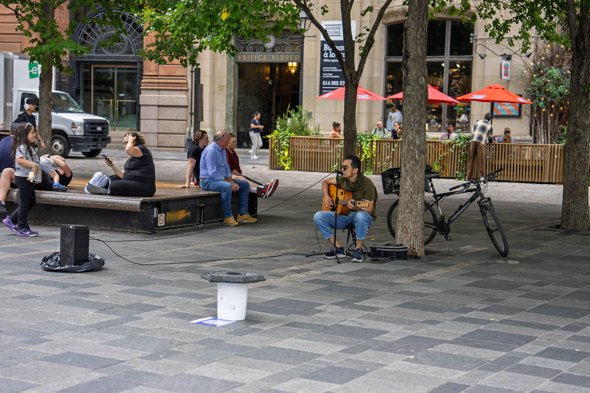 02_septembre_2026Quebec_20266667.jpg — Un guitariste-chanteur assis joue sur la place d'Armes à Montréal, devant l’Édifice Aldred ; des passants assis sur un banc l’écoutent, parasols rouges d’une terrasse à l’arrière-plan.