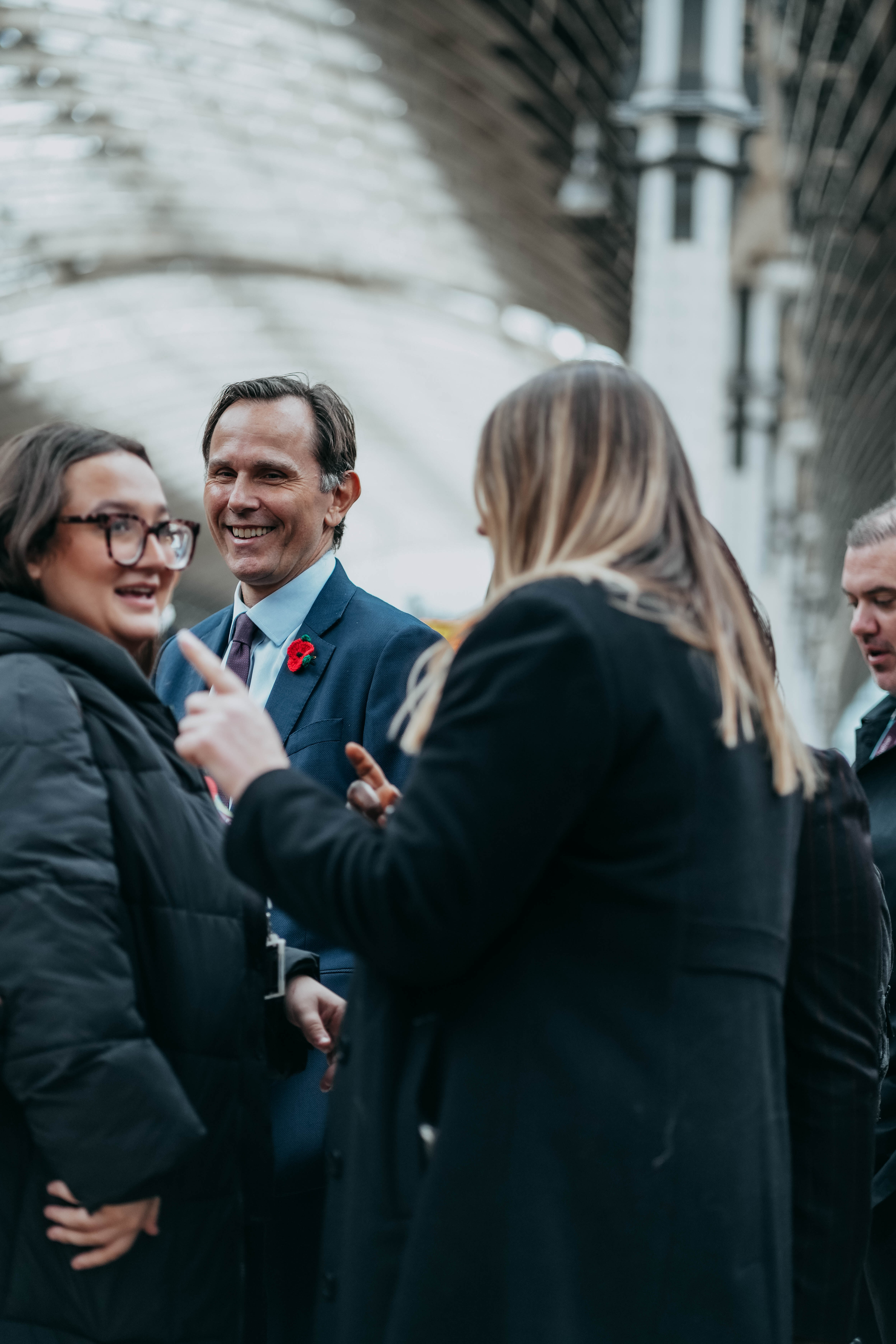 A man in a blue suit with a poppy on his lapel smiles warmly as he listens to a woman during a conversation with a group of people at a train station.