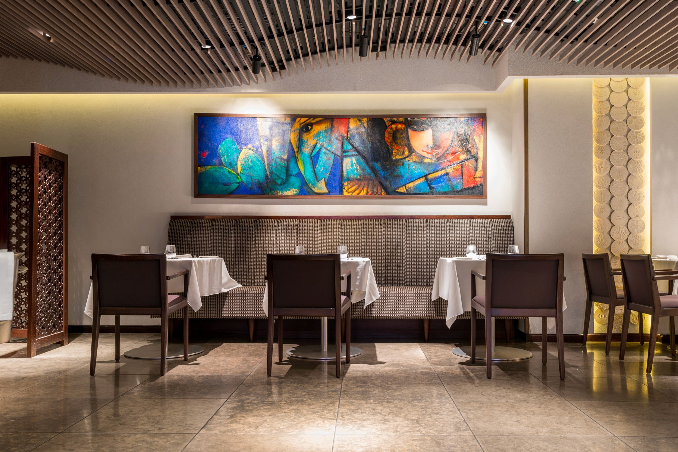 Three tables with white cloths and brown chairs sit in a modern restaurant under a large blue abstract painting.
