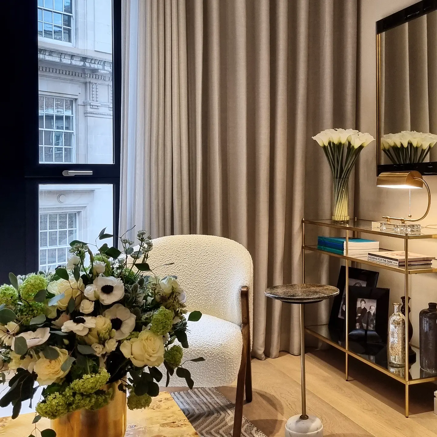 A modern living room features a plush white armchair beside a wooden table adorned with a vibrant floral arrangement, situated next to a tall gold shelf with books and decorative items, framed by a large window with elegant curtains.