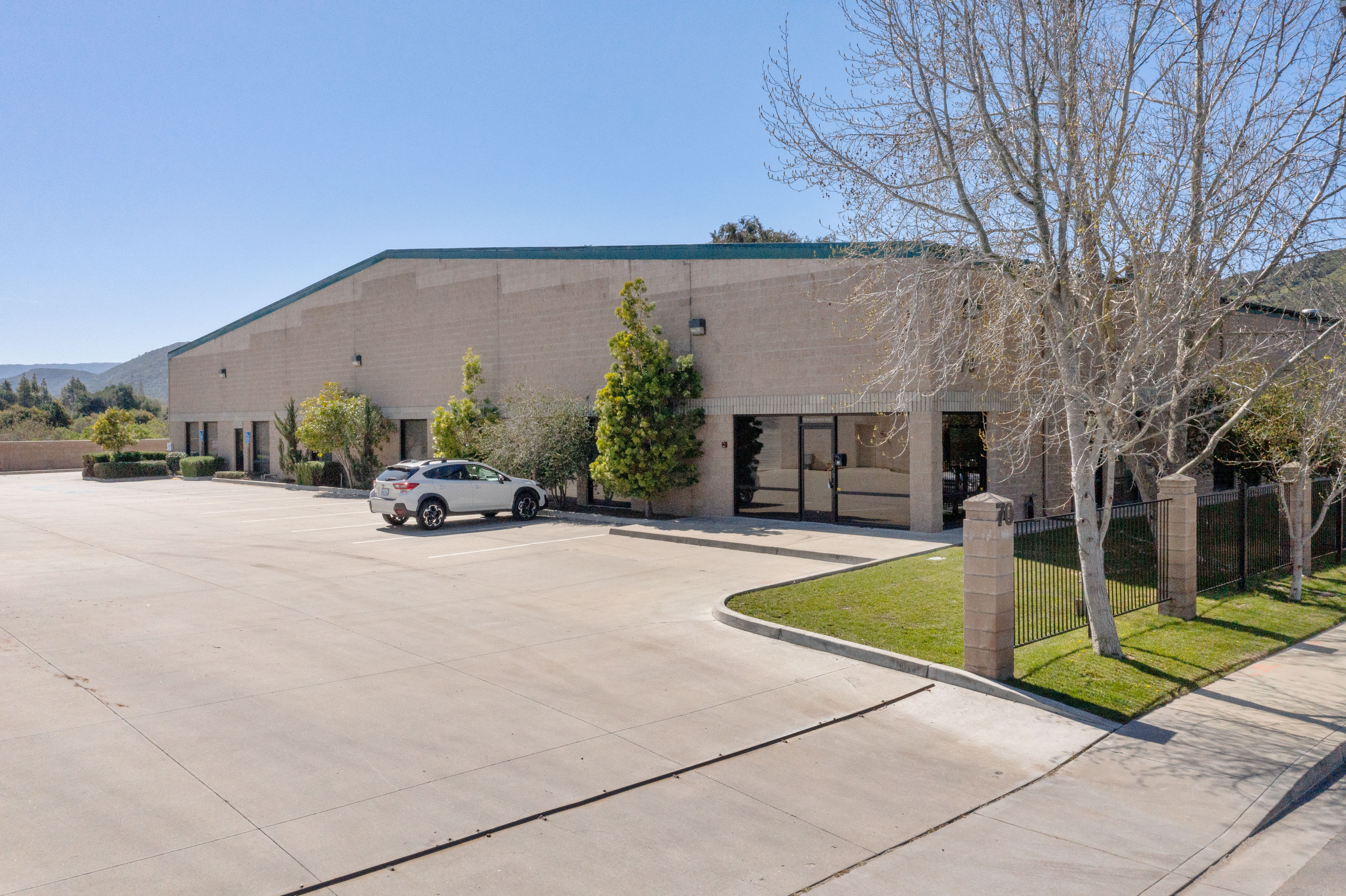 Street-level front exterior of a single-story tilt-up warehouse building with a green roof accent, glass entry doors, mature deciduous trees, a circular drive, and a parked SUV, under a clear blue sky with hills in the distance.