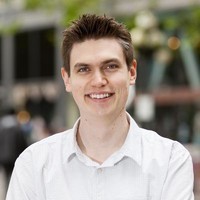A person with short brown hair is smiling while wearing a white shirt, standing outdoors in a city setting with blurred buildings and green trees in the background.