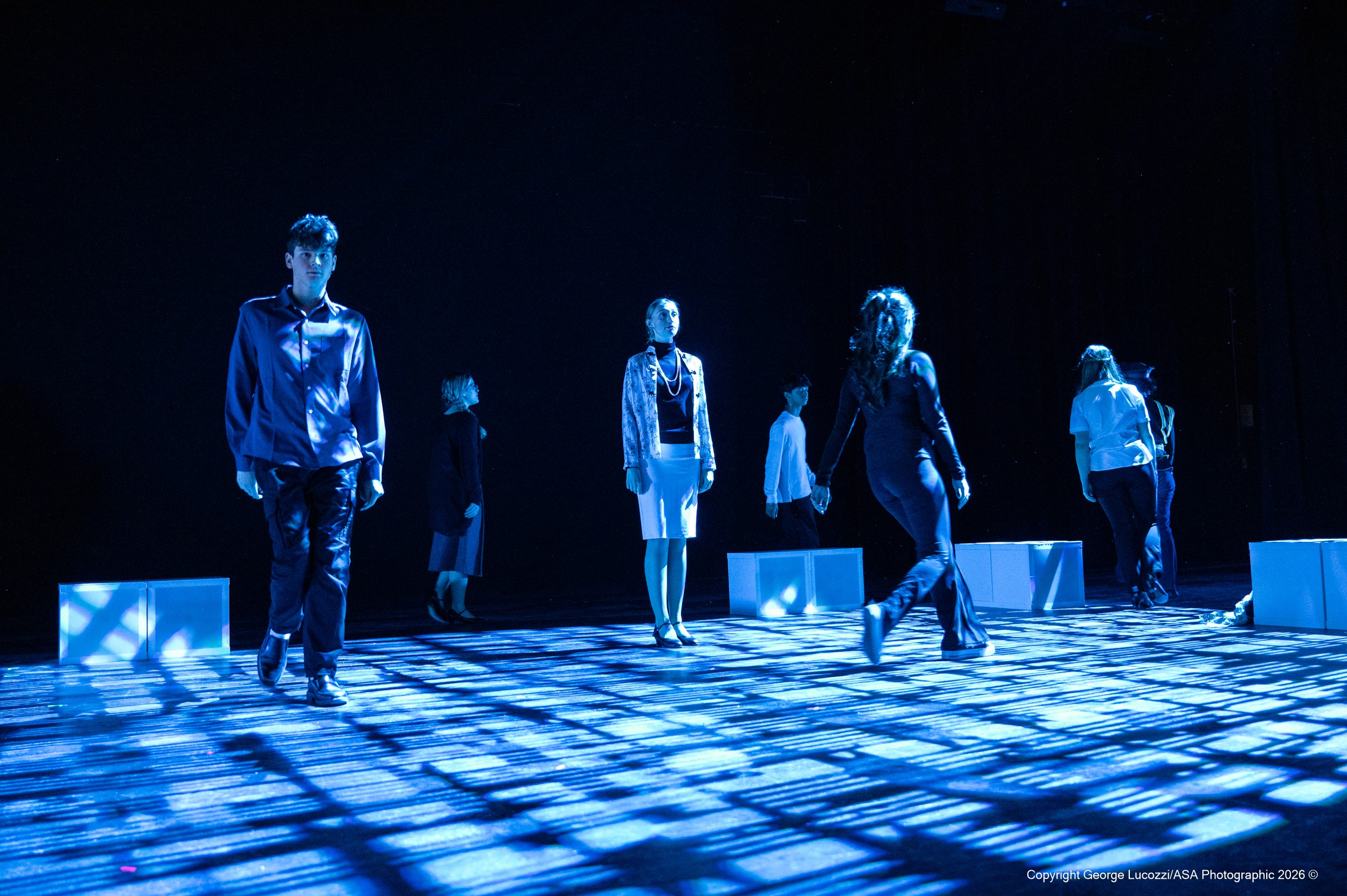 Ensemble members portraying the “Voices” cross the stage in intersecting paths beneath hard-edged blue overhead beams, Rosco Squares (77524) gobos sharply texturing the floor with geometric pattern and movement.