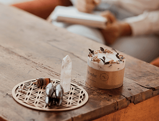 Une photo d'une bougie, d'un cristal blanc et d'un pendule sur un bureau en bois. On peut voir quelqu'un tenir un cahier en fond 