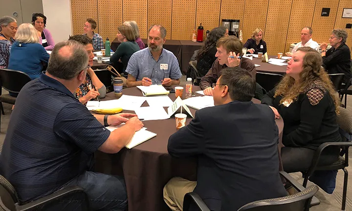 A small group of 6 sitting around a round table with a black table cloth as part of a larger convening. There are papers and pens in front of the attendees.