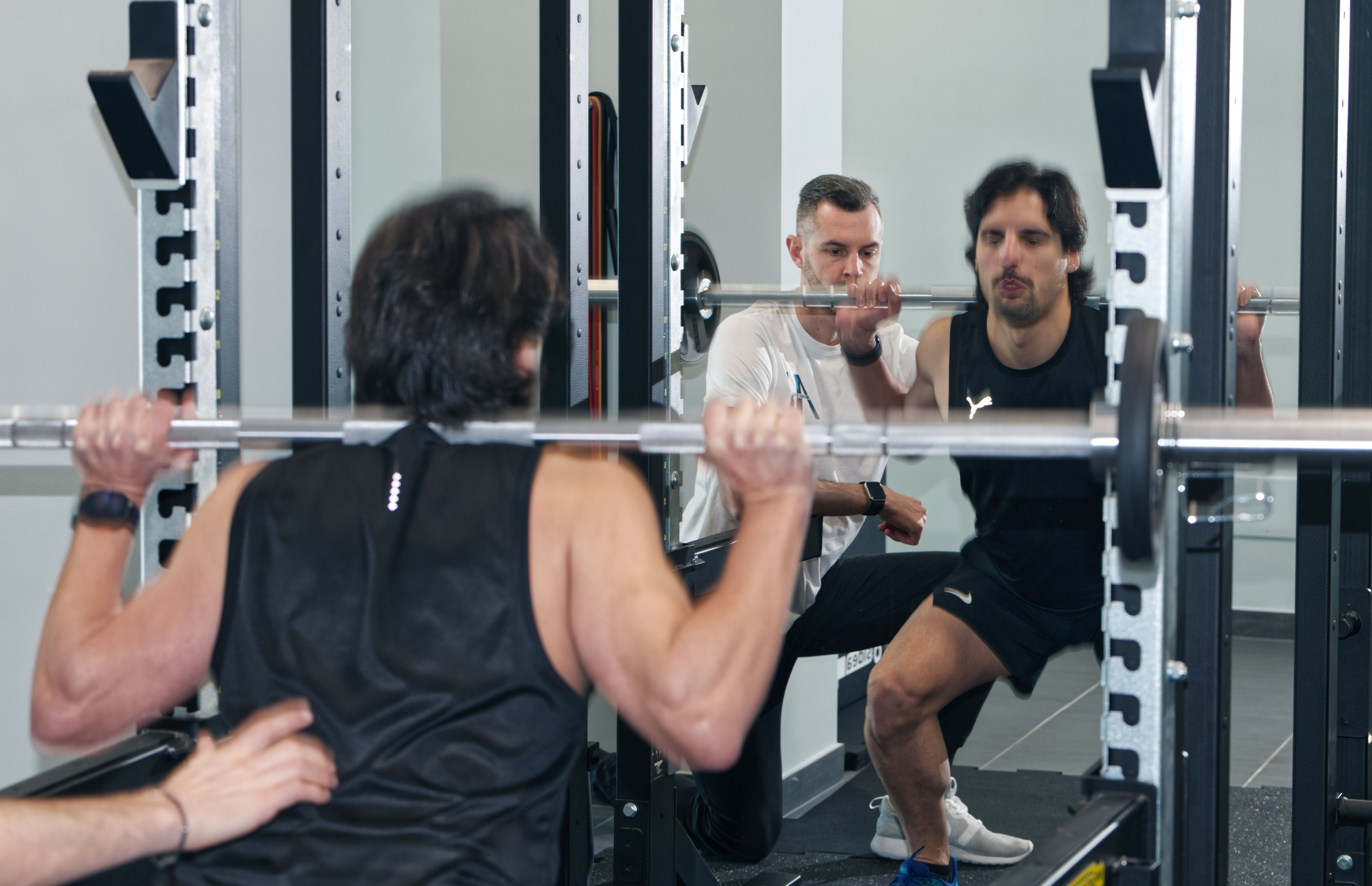 Two men sit at a wooden table with laptops, under a large red lamp. A woman walks by a window. The scene is calm, set against a red-brown wall.