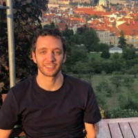 A person is sitting at an outdoor café with a scenic view of a historic European city, featuring red-roofed buildings and greenery, under a clear sky.
