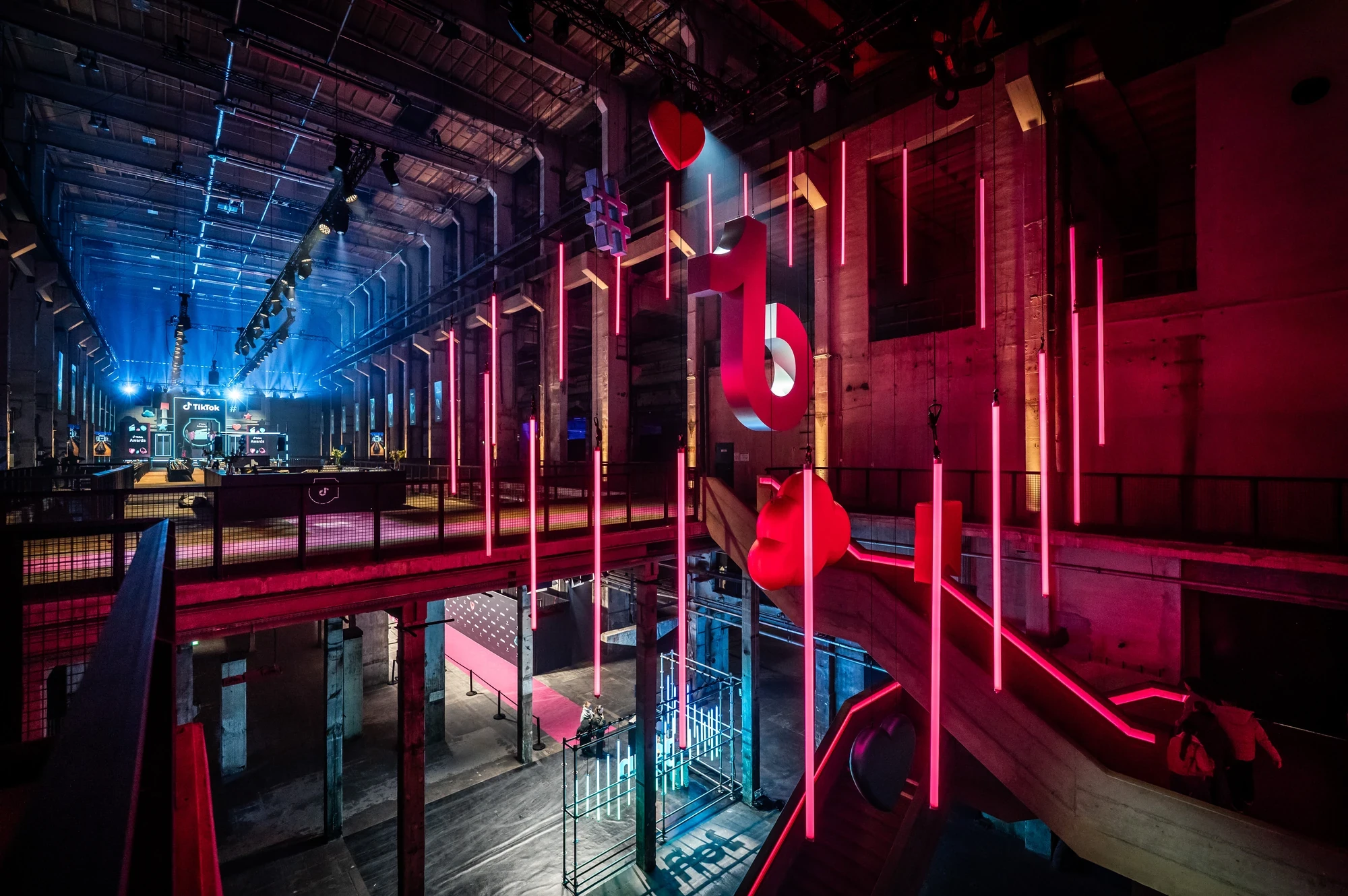 Industrial event hall at the TikTok Awards featuring a suspended light installation of pink neon tubes and floating red logo sculptures.