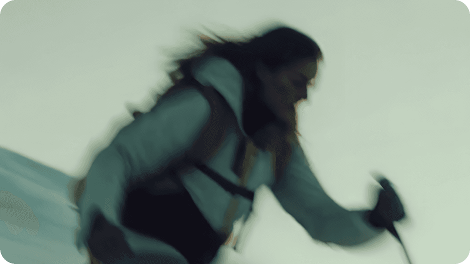 A person in motion is skiing downhill on a snowy slope, wearing a white jacket and holding ski poles, with a blurred background suggesting speed and movement.