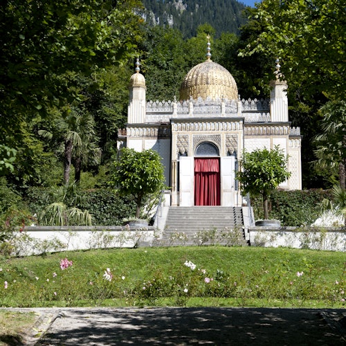 Malá zdobená budova se zlatou kupolí a červeným závěsem, obklopená bujnou zelení a květinami.
