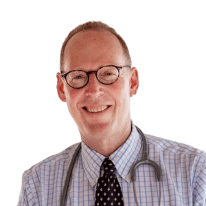 Color portrait of a smiling man wearing glasses, a checkered shirt, and a black polka-dot tie, with a stethoscope draped around his neck. He stands in a covered outdoor walkway, with a blurred background of greenery and buildings, suggesting a medical or humanitarian setting. His friendly expression conveys warmth and approachability.