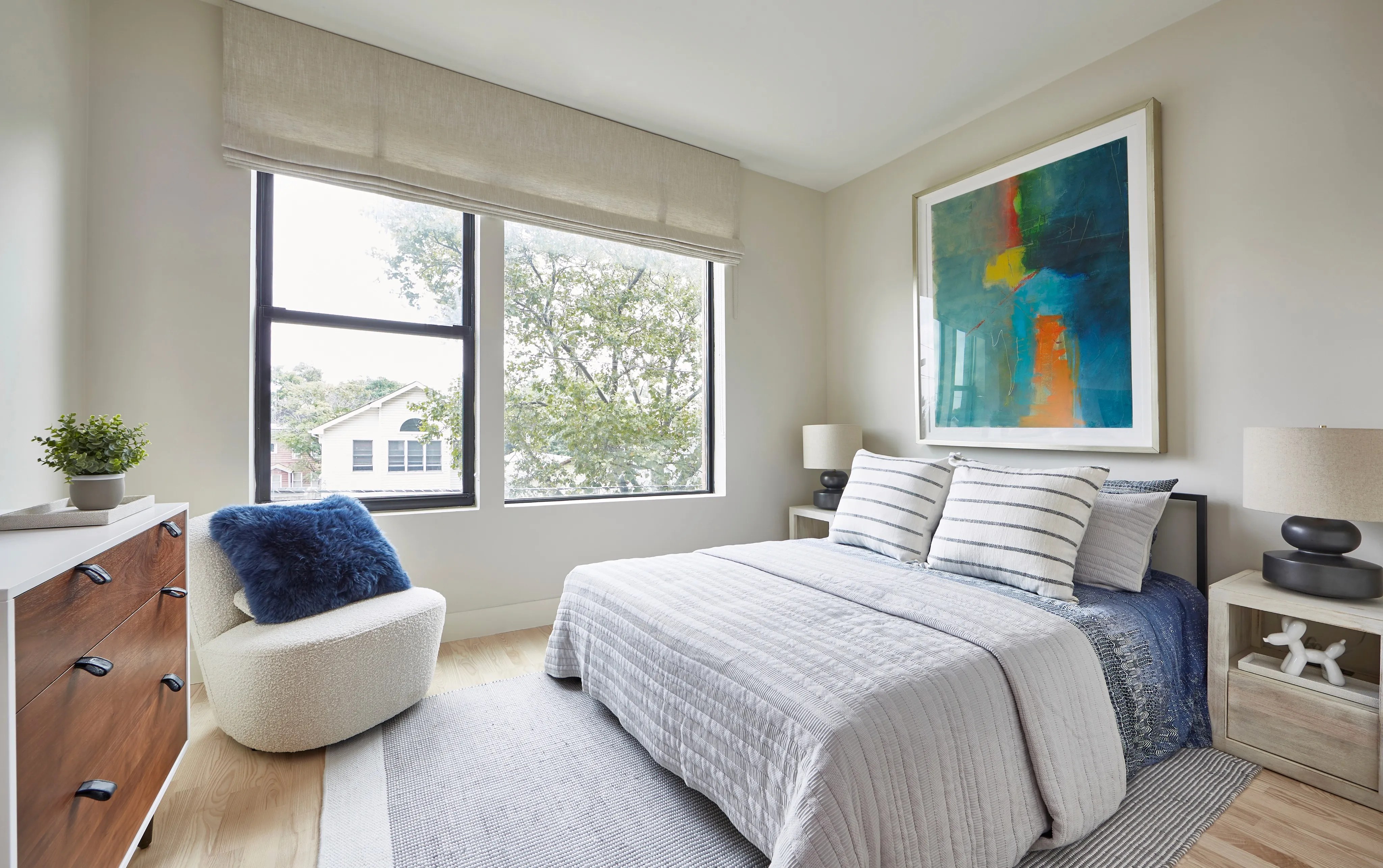 Bright, modern bedroom featuring a double bed, artwork, and large windows. Cozy seating area and wooden furniture.